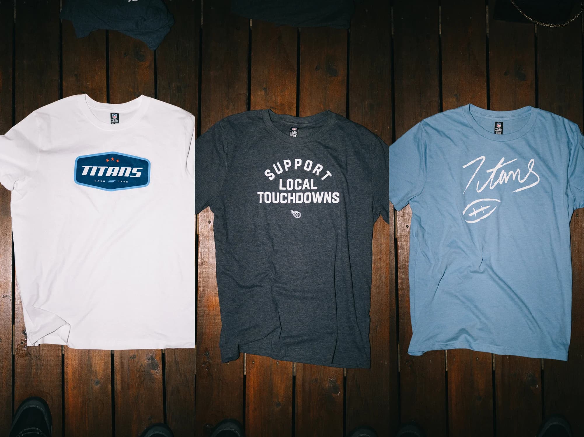 Three Titans tees laid flat on wood: the TITANS badge, Support Local Touchdowns, and the hand-drawn script