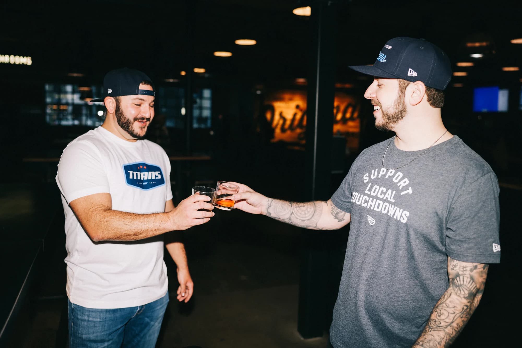Two men in the badge tee and Support Local Touchdowns tee trading a drink at a Nashville bar