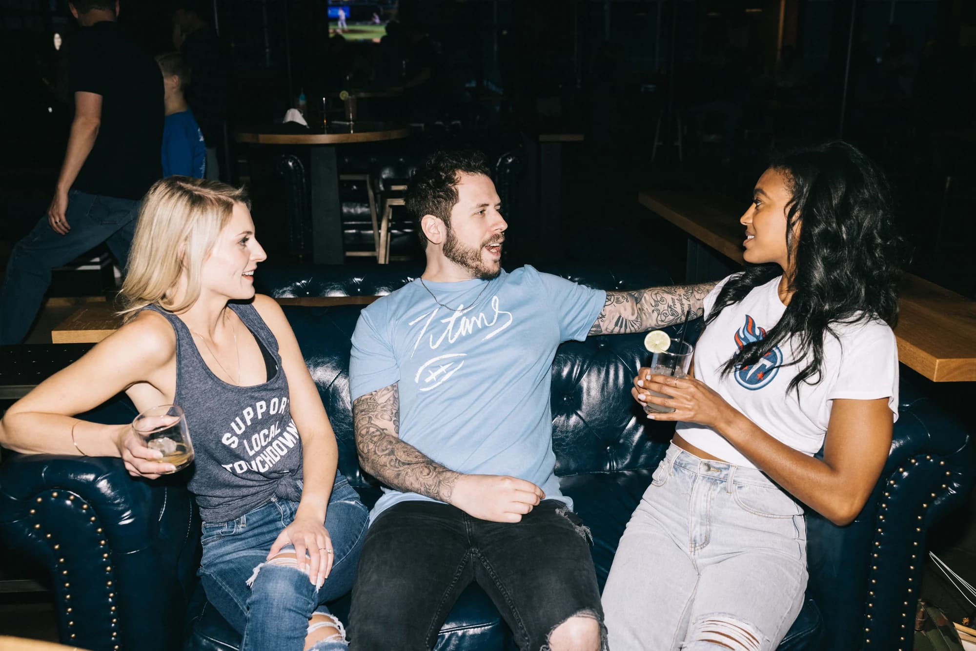Three people on a leather couch wearing the Support Local tank, the script tee and the badge tee